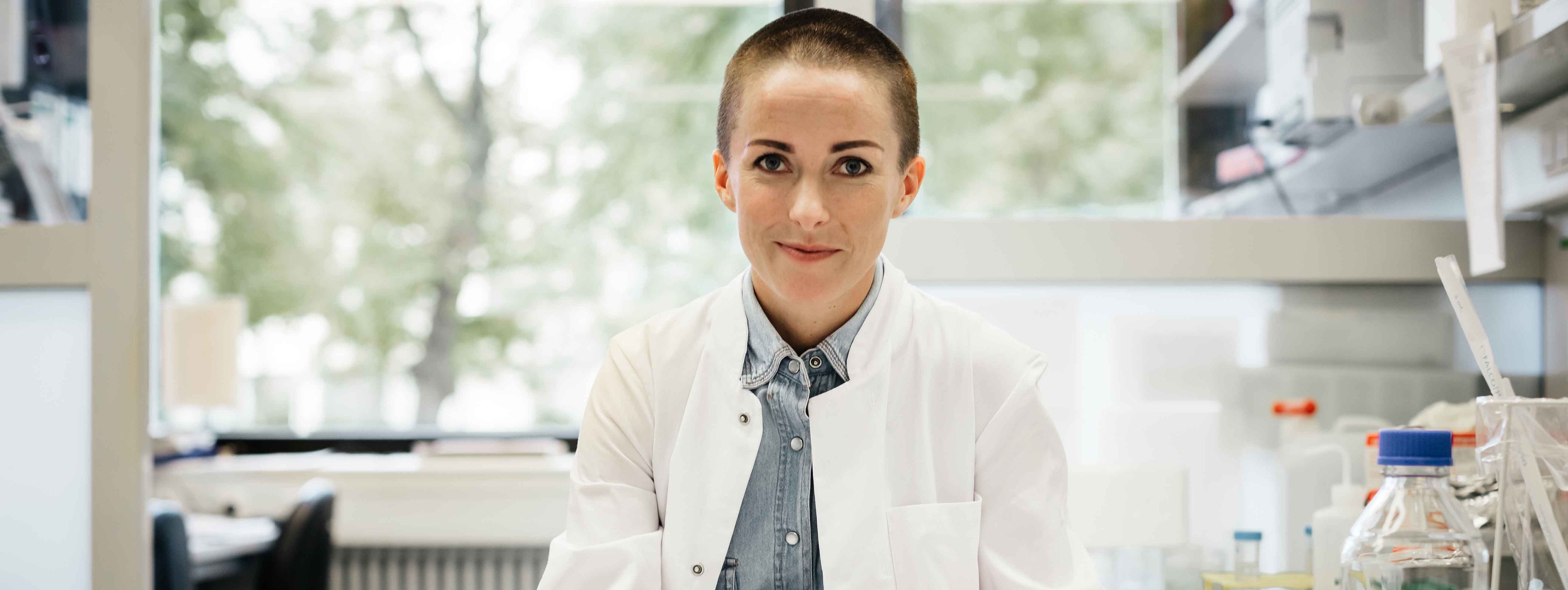 Eine Frau mit Kittel und kurzen Haaren sitzt im Labor und lächelt in die Kamera.