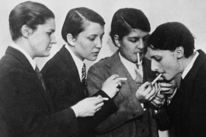 Schwarz-weiß Foto mit jungen Frauen in Anzügen und mit Krawatte, die sich eine Zigarette anzünden.