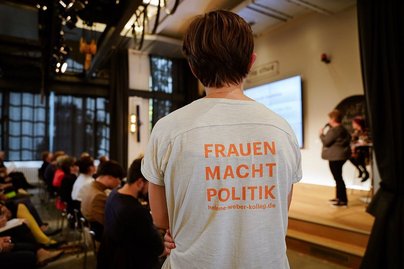 Im Hintergrund Bühne und Publikum. Im Vordergrund eine Person von hinten fotografiert, auf deren Shirt steht: „Frauen Macht Politik. Helene-weber-kolleg.de“.