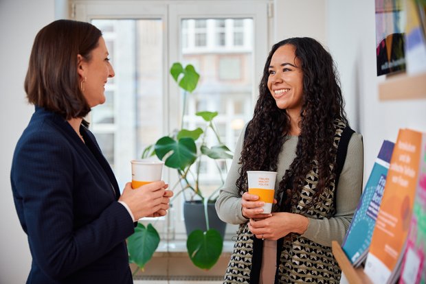 Zu sehen sind zwei Personen die sich unterhalten, beide haben ein Getränk in der Hand. Rechts vorne sind einige Broschüren zu sehen. Im Hintergrund steht eine Pflanze auf einer Fensterbank.